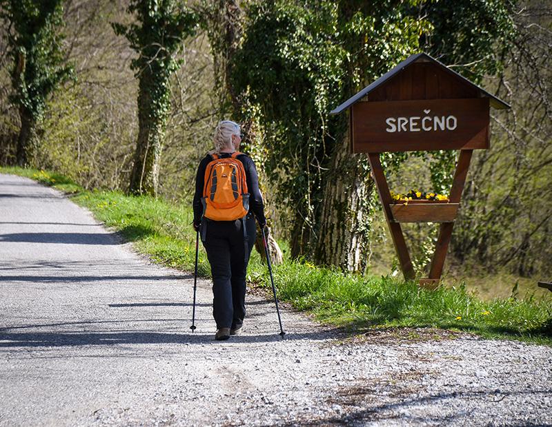 Woman walking with poles past a welcome sign in a Slovenian village, part of a self-guided countryside hiking holiday.